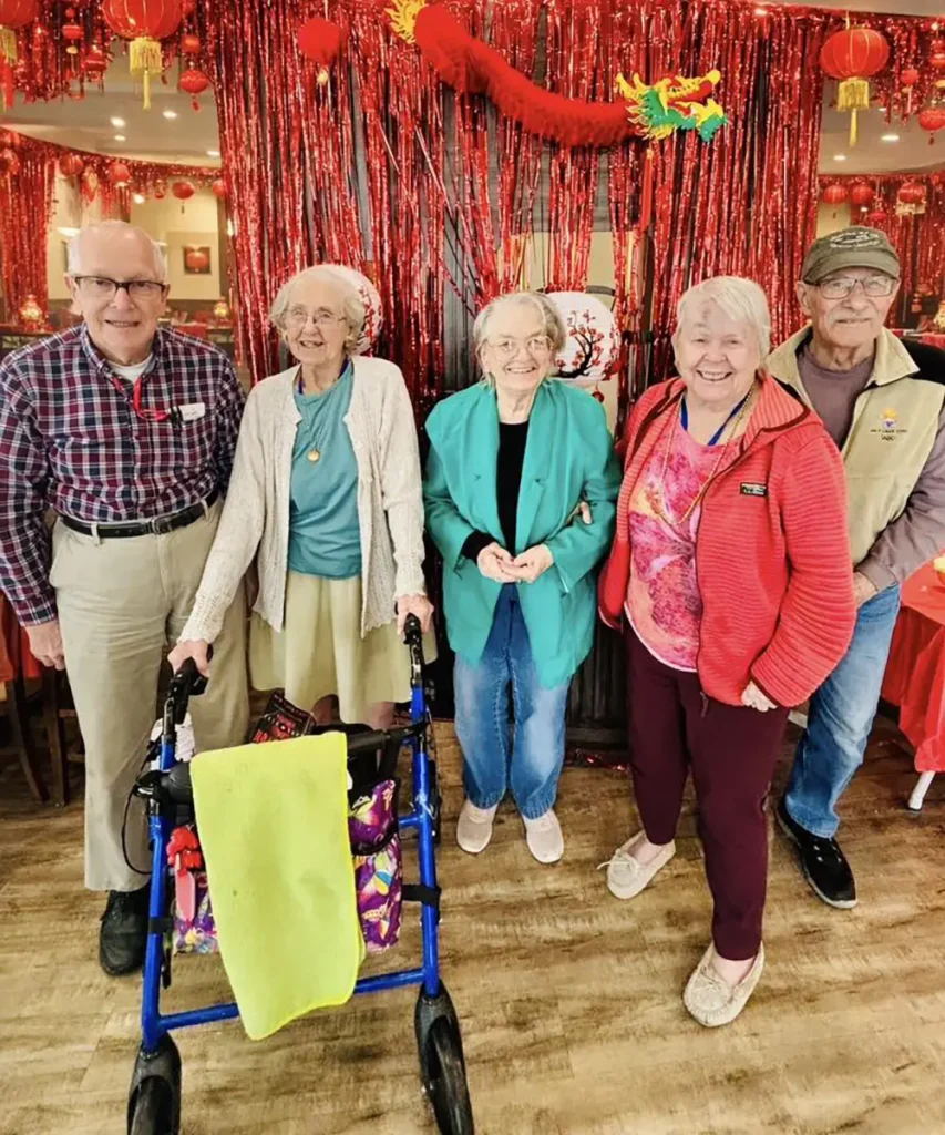 Five older adults stand indoors with red decorations and a walker visible, smiling for a group photo as they celebrate the Lunar New Year.