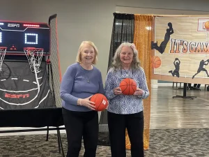 Two older women stand indoors holding basketballs, smiling in front of an indoor basketball game setup and a sports-themed banner, enjoying some March Madness fun.