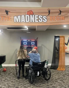 A person in a wheelchair plays an arcade basketball game with assistance from another person under a banner reading "The Madness Begin," capturing the spirit of March Madness Fun.