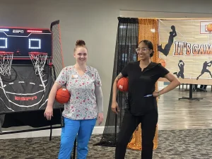 Two women, one in medical scrubs and one in black attire, stand with basketballs near an indoor arcade game and a sports-themed backdrop, ready to join the March Madness fun.