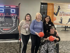 Four women, one in a wheelchair, pose with small basketballs in front of an indoor basketball game and a sports-themed banner, ready to enjoy some March Madness fun.