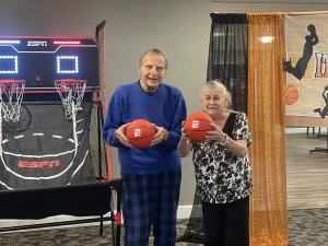 Two older adults stand indoors holding basketballs in front of a basketball arcade game, smiling for the camera and enjoying some March Madness Fun.