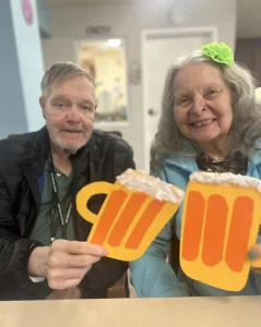 An elderly man and woman sit side by side indoors, each holding a large paper cutout of a beer mug with foam, celebrating National Beer Month together.
