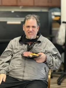 A man wearing a gray jacket sits indoors and smiles at the camera while holding a plate of assorted food in his lap, celebrating National Beer Month.