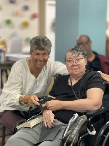 Two older women pose together indoors; one is seated in a wheelchair holding a book, while the other stands beside her, smiling with a hand on her shoulder as they celebrate National Beer Month.