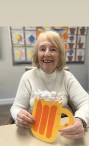 An older woman in a white sweater smiles and holds a handmade craft shaped like a mug of beer, celebrating National Beer Month in a room with children's artwork displayed on the wall behind her.
