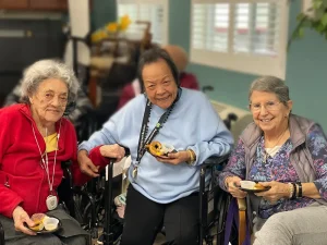 Three elderly women sit together indoors, smiling and holding snacks in bowls while celebrating National Beer Month. Two are in wheelchairs, and all are wearing glasses and casual clothing.