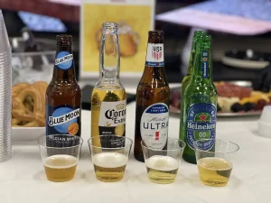 Four different beer bottles—Blue Moon, Corona Extra, Michelob Ultra, and Heineken 0.0—stand behind four small cups of poured beer on a table with snacks in the background, making it a perfect setup to celebrate National Beer Month.