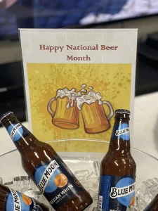 Two bottles of Blue Moon beer on ice sit in front of a sign with two beer mugs that reads "Happy National Beer Month." Cheers to celebrating National Beer Month in style!.