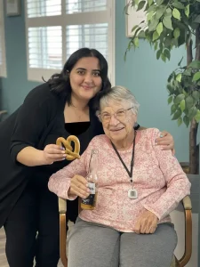 Two women pose and smile for National Beer Month; one sits holding a bottle of beer, while the other stands behind her with a pretzel. They are indoors near a potted plant.