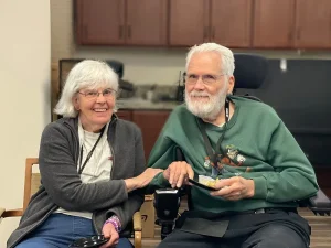 An older woman and man sit together indoors. The woman smiles while holding the man’s arm; the man, in a wheelchair, holds a plate of food and smiles, enjoying each other's company during National Beer Month. Cabinets are visible in the background.