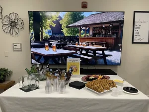 A table with bottled drinks, empty glasses, pretzels, and snacks is set up in front of a TV displaying an outdoor beer garden scene—perfect for celebrating National Beer Month.