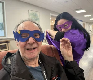An older man and a woman wear purple superhero masks and capes indoors, smiling and posing for the camera in celebration of National Superhero Day.