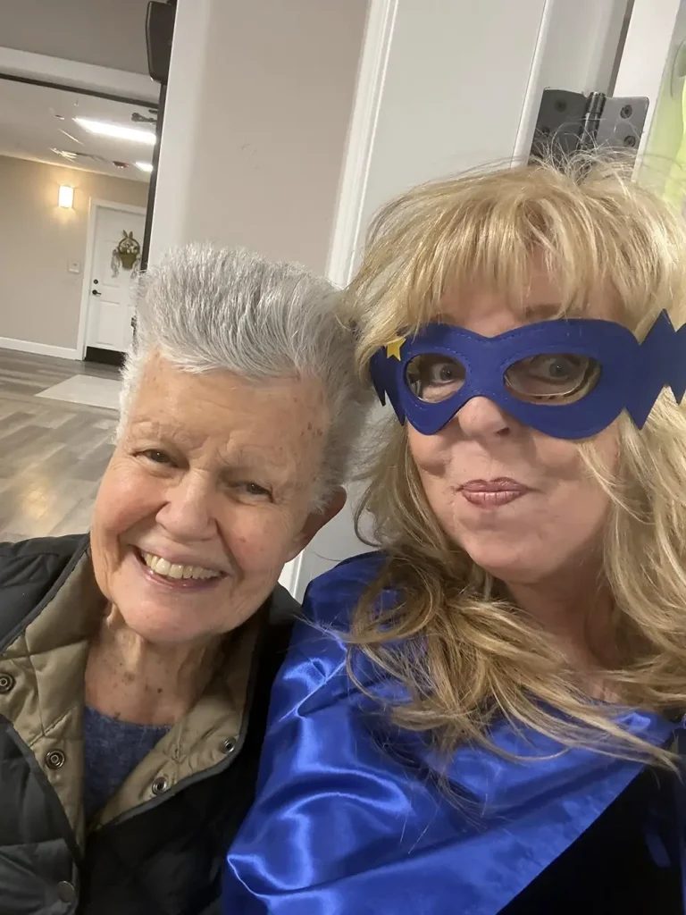 Two women pose together indoors; one is smiling while the other wears a blue cape and mask, resembling a superhero costume&mdash;perfect for celebrating National Superhero Day.