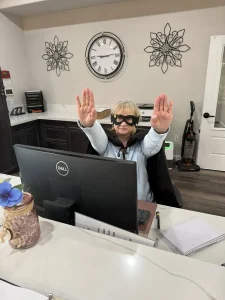 A person in an office celebrates National Superhero Day, wearing a cape and sunglasses behind a desk with a computer, striking a playful pose with both hands raised.
