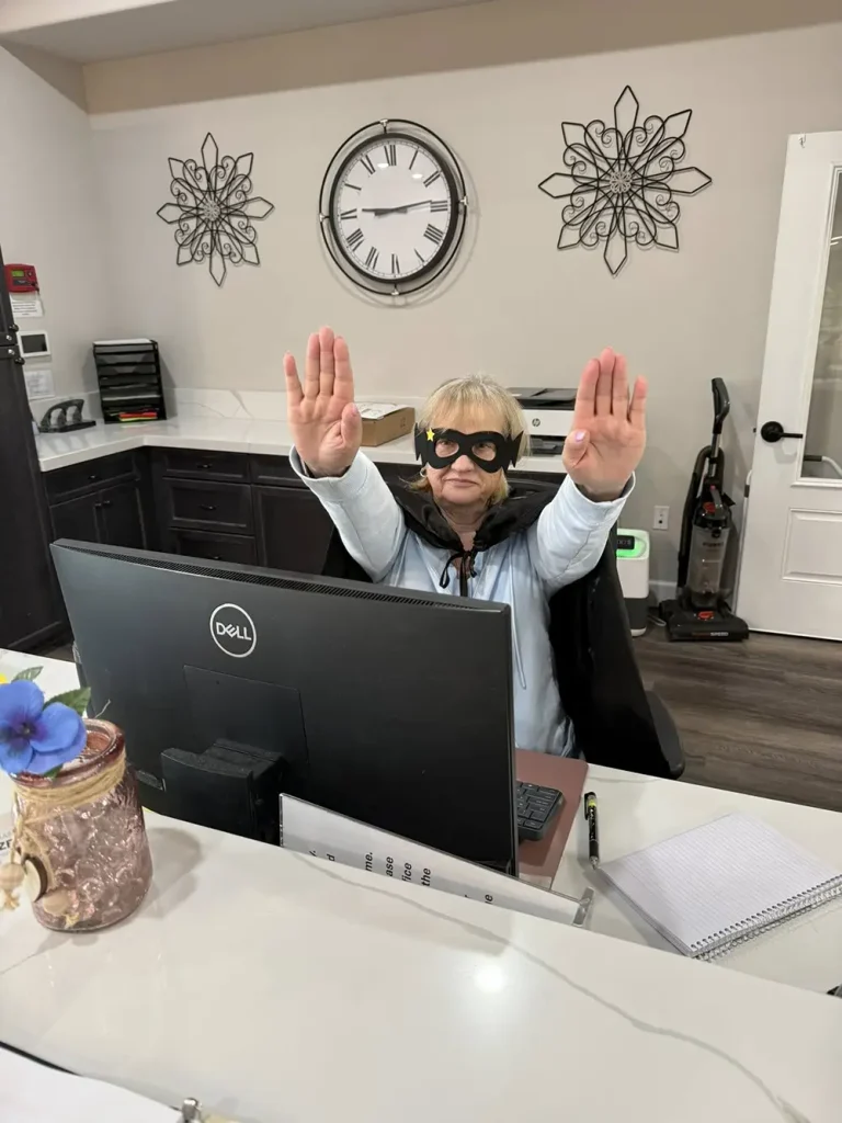 A person in an office celebrates National Superhero Day, wearing a cape and sunglasses behind a desk with a computer, striking a playful pose with both hands raised.