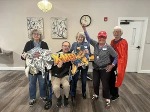 Five older adults pose indoors for National Superhero Day; one holds an X-Men sign, another wears a red cape. Art hangs on the wall behind them, with a clock and door in the background.