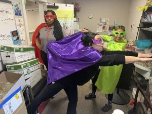 Three people in colorful superhero capes and masks pose playfully in an office surrounded by boxes and office supplies, celebrating National Superhero Day with cheerful energy.
