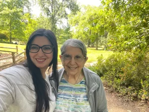 Two women, one younger and one older, stand outdoors in a park, smiling at the camera with green trees and bushes in the background, celebrating National Garden Meditation Day.