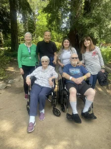 Six adults, including two in wheelchairs, pose together outdoors on a sunny day with trees and greenery in the background, celebrating National Garden Meditation Day and embracing garden mindfulness.