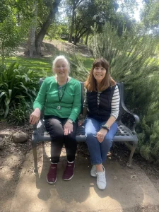 Two women sit side by side on a metal bench in a park, surrounded by greenery and trees, smiling at the camera as they embrace Garden Mindfulness.