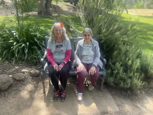 Two elderly women sit side by side on a bench outdoors, enjoying garden mindfulness amid greenery and sunlight—perfect serenity on National Garden Meditation Day.