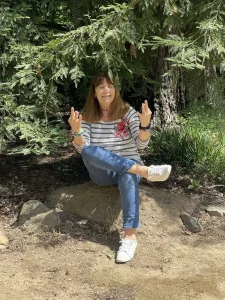 A woman sits on a rock under tree branches, smiling with mindfulness outdoors. With legs crossed and hands raised, she enjoys the calm of a meditation garden in her striped floral shirt, blue jeans, and white sneakers.