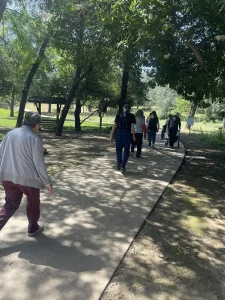 A group of people walks along a shaded concrete path in a park with trees and greenery, enjoying mindfulness on a sunny day—perhaps celebrating National Garden Meditation Day.