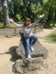 A woman practices garden mindfulness, sitting on a large rock with raised arms and a smile, surrounded by trees and a wooden fence on a sunny day.