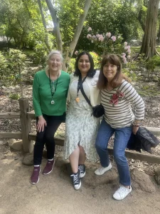 Three women pose and smile together in a garden setting, enjoying the sunlight, trees, and flowers—capturing the spirit of garden mindfulness and serenity.