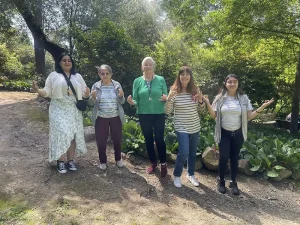 Five women stand outdoors in a garden, facing the camera with trees and greenery in the background. Celebrating National Garden Meditation Day, they hold their hands up and smile, embracing garden mindfulness together.