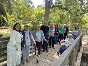 A group of older adults and caregivers stand and sit together on a wooden bridge in a park, surrounded by trees and greenery, enjoying garden mindfulness in celebration of National Garden Meditation Day.