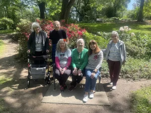 Six older adults pose for a group photo outdoors in a garden, surrounded by blooming flowers and greenery, celebrating National Garden Meditation Day and embracing the mindfulness that meditation in gardens can bring.