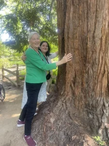 Two women stand outdoors, smiling and hugging a large tree trunk, celebrating garden mindfulness. A wooden fence and lush greenery suggest a peaceful spot for meditation—perfect for National Garden Meditation Day.