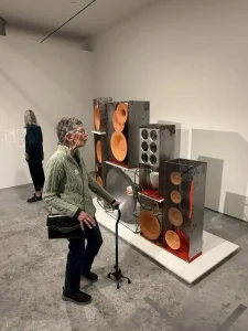 An elderly person with a cane enjoys an art outing, viewing large, rusted metal speakers displayed on a platform in the Nevada Museum of Art gallery. Another visitor reads a wall label in the background.