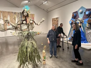 Three people enjoy an art outing at the Nevada Museum of Art, observing a large, nature-inspired humanoid sculpture surrounded by colorful artworks on the gallery walls.