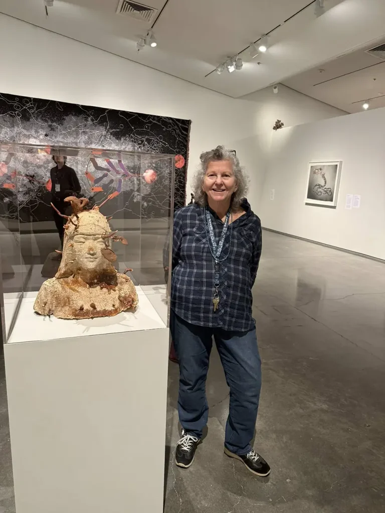 A woman stands next to a sculpted bust displayed on a pedestal during an Art Outing at the Nevada Museum of Art, with vibrant artworks visible on the surrounding gallery walls.