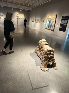 During an art outing at the Nevada Museum of Art, a woman observes a large, animal-like sculpture made of stacked materials in the gallery, surrounded by paintings and another visitor in the background.