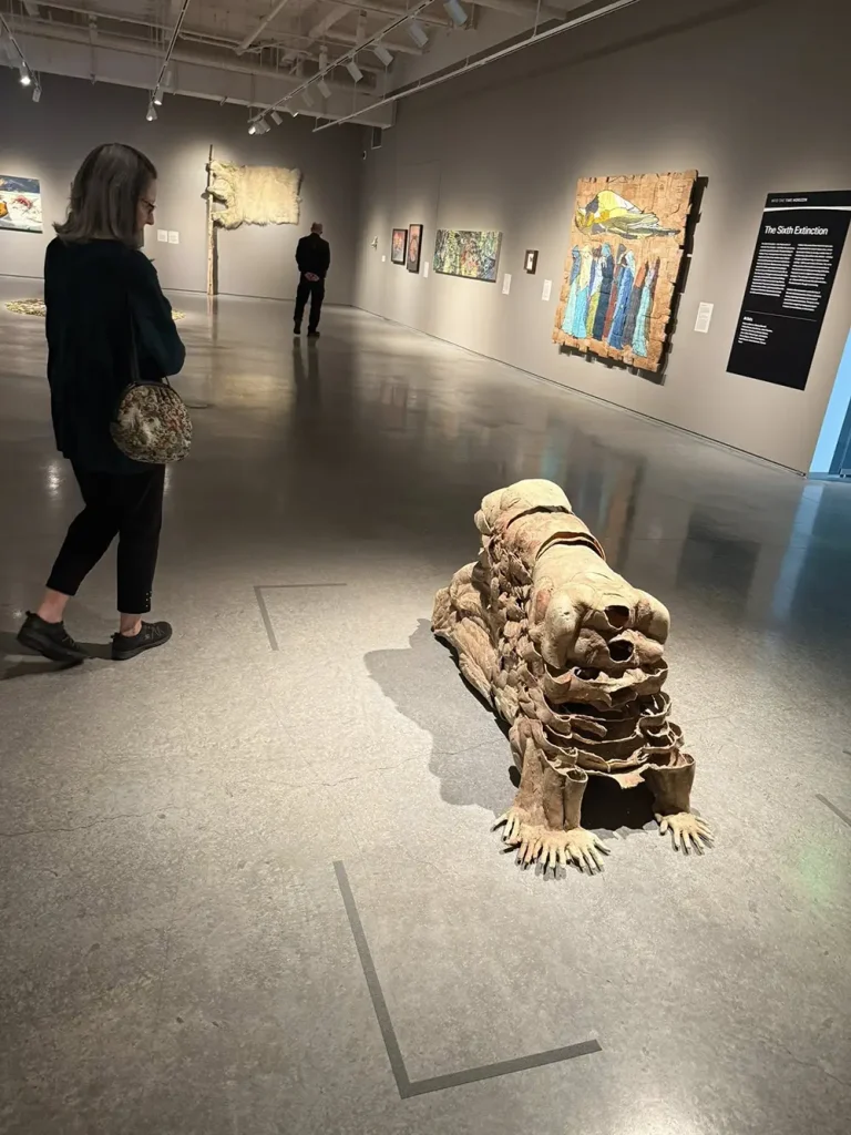 During an art outing at the Nevada Museum of Art, a woman observes a large, animal-like sculpture made of stacked materials in the gallery, surrounded by paintings and another visitor in the background.