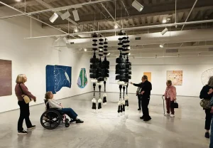 People enjoy an art outing at the Nevada Museum of Art, where a woman in a wheelchair and others stand, discussing abstract paintings and hanging sculptures displayed throughout the gallery.