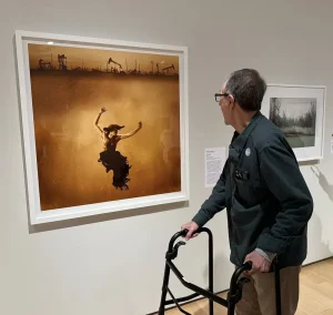 An older man with a walker looks at a framed photograph of a person submerged in water with oil pumps in the background, displayed on a Nevada Museum of Art gallery wall during a museum outing.