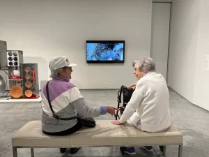 Two elderly women sit on a bench in the Nevada Museum of Art, talking. A video screen displaying abstract art is mounted on the wall in front of them.