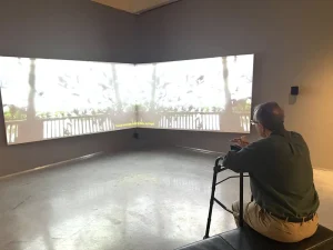 An older man with a walker enjoys a museum outing, watching a large, corner-mounted projection screen with bright scenes and yellow subtitles in the dimly lit Nevada Museum of Art.