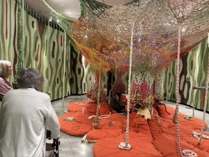 Two people enjoy an art outing as they observe a colorful, web-like yarn installation suspended above orange cushions in a gallery at the Nevada Museum of Art, with patterned green and brown walls enhancing the creative space.