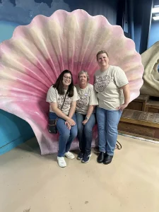 Three women in matching beige t-shirts and jeans pose in front of a large pink seashell display indoors, capturing a fun outing at the Norcal Aquarium.