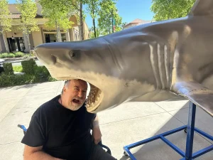 A man poses with his head inside the open mouth of a large shark statue outdoors at the Norcal Aquarium on a sunny day, capturing a playful moment during his wildlife adventures.