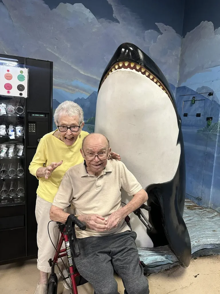 An elderly woman and man, with the man seated in a wheelchair, smile during their Norcal Aquarium outing, posing in front of a large orca statue next to a vending machine.