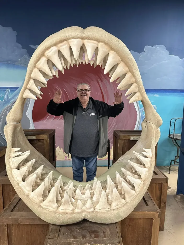 A man stands inside a large replica of a shark jaw, smiling and raising his hands during a fun outing at the Norcal Aquarium, with a painted ocean background behind him.