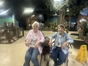 Two elderly women sit in wheelchairs inside a Norcal wildlife exhibit with animal enclosures and trees; a "Caution Wet Floor" sign is visible on the right, adding to their indoor Wildlife Adventures.