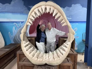 Two smiling older adults pose and wave inside a large replica of a shark's open jaws at the Norcal Aquarium, surrounded by painted ocean-themed walls for an unforgettable Wildlife Adventures experience.